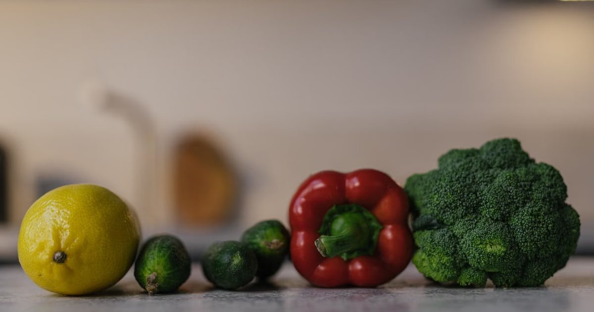 Close-up of colorful fresh produce on a kitchen counter, featuring lemon, cucumbers, red bell pepper, and broccoli.