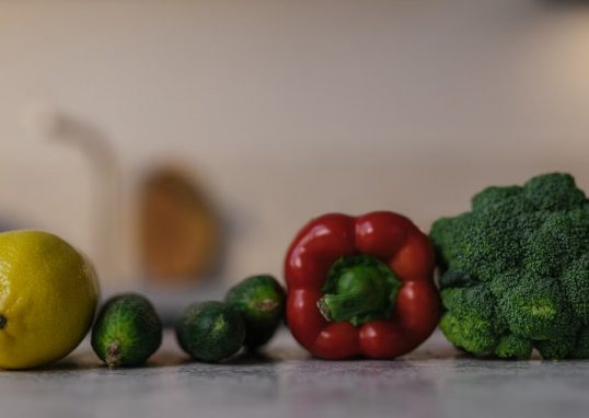 Close-up of colorful fresh produce on a kitchen counter, featuring lemon, cucumbers, red bell pepper, and broccoli.