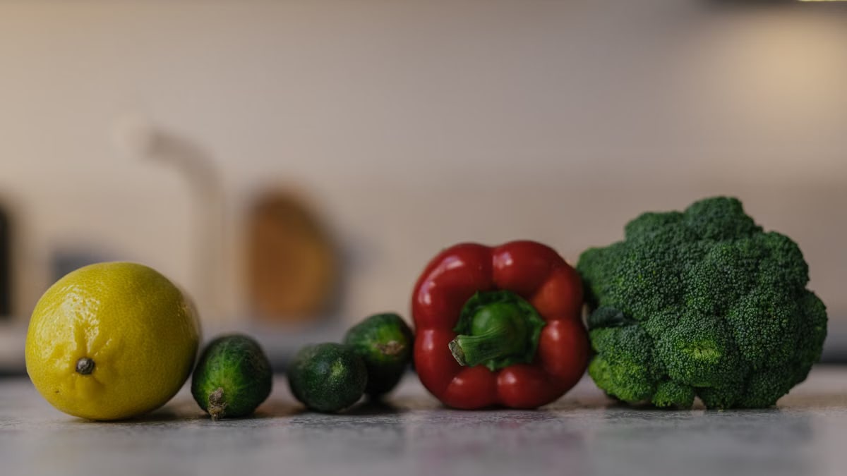 Close-up of colorful fresh produce on a kitchen counter, featuring lemon, cucumbers, red bell pepper, and broccoli.