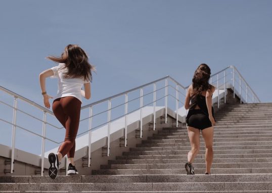 Two women running up outdoor stairs for fitness under a clear blue sky.